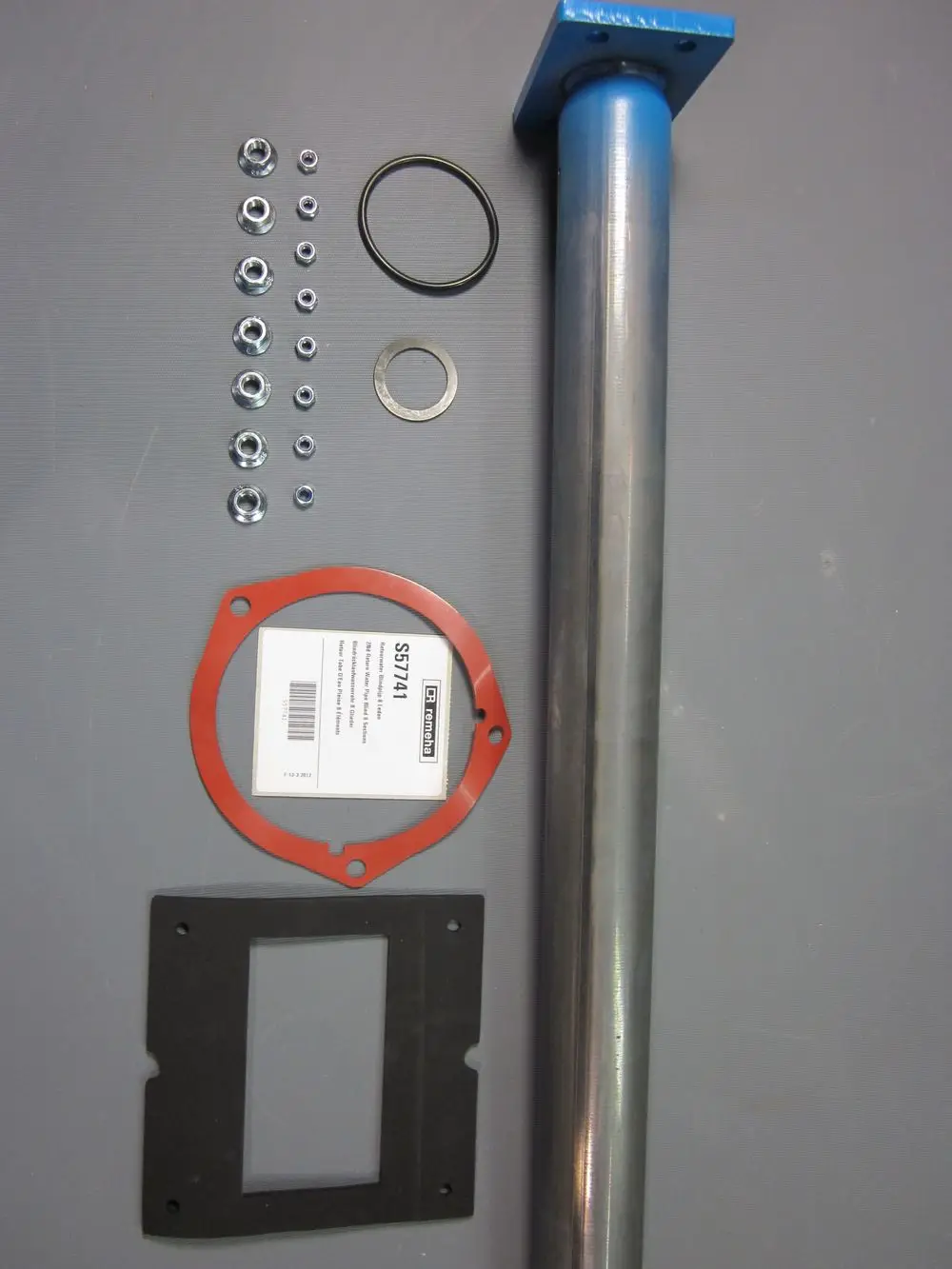 Remeha Zubehörteile, einschließlich eines Metallrohrs, Dichtungen, Unterlegscheiben und Schrauben, auf einem grauen Untergrund angeordnet.
